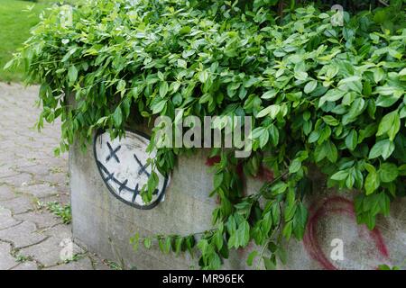 Graffiti di un viso Smiley con X occhi e bocca cucita. La superficie si trova sul lato di una piantatrice di calcestruzzo con foglie che fuoriescono sul bordo. Foto Stock