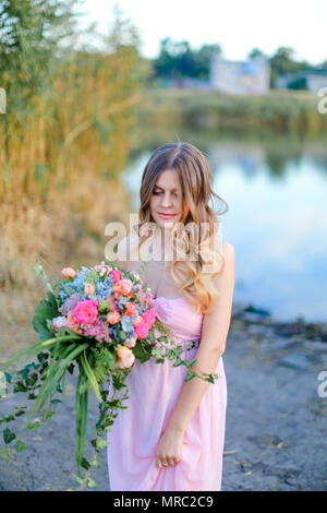 Femmine gravide di persona che indossa l'abito rosa con bouquet di fiori in piedi vicino al lago. Foto Stock