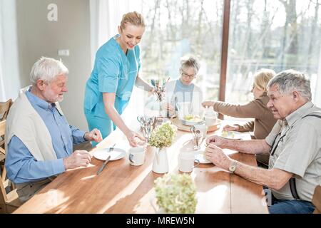 Femmina lavoratore di cura che serve adulti senior al tavolo per la cena in casa di cura. Foto Stock