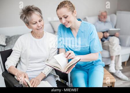 Lavoratore di cura di libro di lettura con il senior donna nella casa di cura. Foto Stock