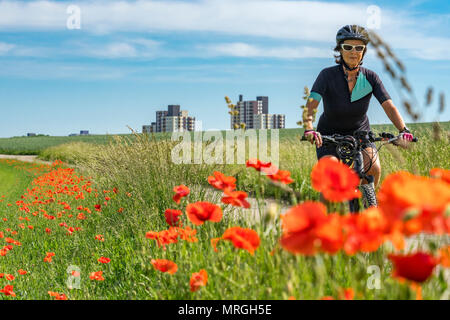 Bella donna Senior in sella a una e-Mountainbike nel sobborgo di una grande città, circondata da verdi campi e papavero rosso Foto Stock