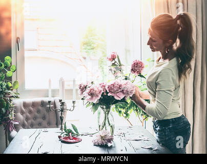 Bella donna moderna con pony tail hair disponendo le peonie mazzetto in vaso di vetro sul tavolo in vetro in salotto. Stile di vita. Casa felice Foto Stock