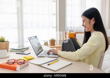 Femmina asiatica di sedersi con le ginocchia fino utilizzare i social media con il computer portatile sul tavolo e bere il succo di arancia in camera da letto a casa.Il lavoro a casa concetto.lavoro da casa.rel Foto Stock