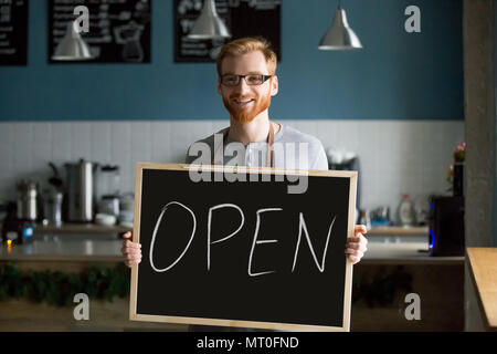 Redhead uomo sorridente tenendo aperte segno invitante per nuovo cafe Foto Stock