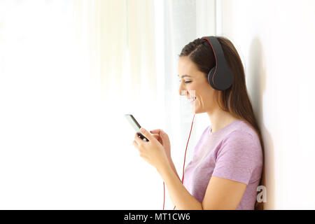 Profilo di un felice teen vestita di nero le cuffie per ascoltare musica selezionando i brani in uno smart phone a casa Foto Stock