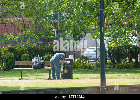 Un uomo perquisisce un bidone in un parco pubblico in una giornata di sole, evidenziando la povertà urbana e i problemi sociali in un ambiente urbano, nel Regno Unito Foto Stock