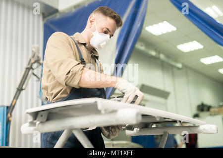 Basso angolo vista del giovane maschio auto tecnico nella maschera protettiva la lucidatura auto dettaglio con la macchina di rettifica in officina di riparazione Foto Stock