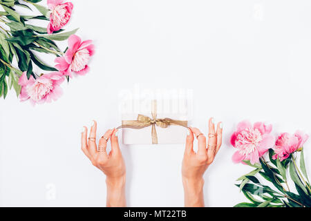 La donna apre il suo presente, vista dall'alto. La femmina le mani si snoda golden nastro sulla confezione regalo tra la peonia rosa fiori, festosa piana stilizzazione dei laici. Foto Stock