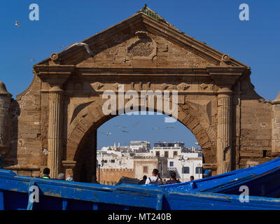 Antico arco di pietra sul molo del porto della città di Essaouira, nel visibile a distanza di edifici bianchi della medina, contro il cielo blu di sfondo, Mo Foto Stock