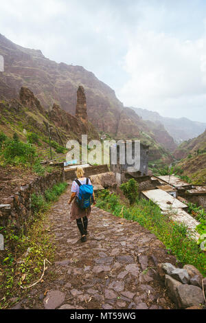 Ragazza con zaino camminando verso il basso lungo il percorso di trekking per la verdeggiante valle Xo-Xo. Picchi di montagna e il villaggio locale è visibile nella parte anteriore. Santo Antao Isola Capo Verde Foto Stock