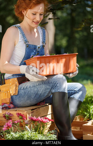 Giovane donna in salopette da wellingtons e seduta su una gabbia in legno all'esterno, nel cortile e tenendo un vaso di fiori Foto Stock