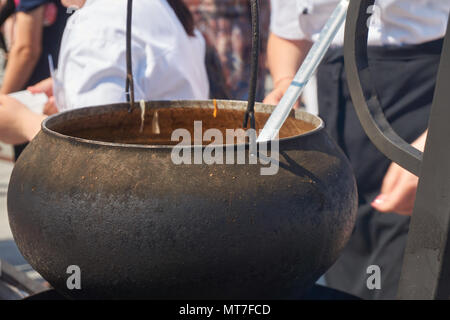 Lo chef con un grembiule nero un vecchio metallo colato pentola in ferro o vaso sospeso da un telaio in una vista ravvicinata Foto Stock