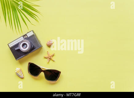 Traveler accessori: verde tropicale di foglie di palma, fotocamera, conchiglie e gli occhiali da sole sono su sfondo giallo con copy-spazio. Vista dall'alto di viaggio o va Foto Stock