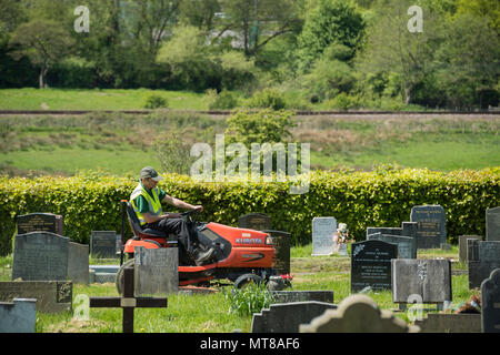 Uomo al lavoro (dipendente tenendo conto del consiglio locale) si siede sul giro sul tosaerba e tagli di erba tra lapidi - Cimitero Guiseley, West Yorkshire, Inghilterra, Regno Unito. Foto Stock