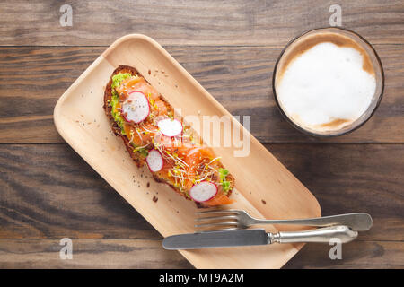 Pane rustico tostato con purea di avocado, salmone affumicato, Rafano, broccoletti e semi servita in un piatto di legno con un caffè con latte in un rustico Foto Stock