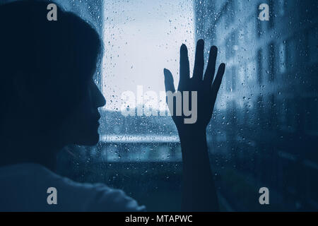 Triste lonely depressione umore silhouette di donna toccare le finestre di vetro scuro piovosa giornata fuori Foto Stock