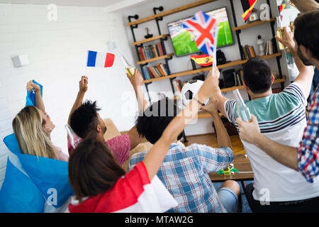 Gli amici con un drink guardando la partita di calcio sulla TV di casa e celebrando la vittoria Foto Stock