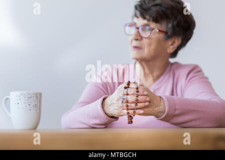 Foto con sfondo sfocato di sambuco donna tenendo un rosario e seduti a tavola con la tazza di tè Foto Stock