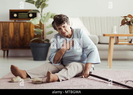 Disabilitato il sambuco signora seduta sul pavimento con un bastone da passeggio e tenendo premuto il suo torace dopo la caduta verso il basso Foto Stock