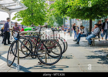 Biciclette parcheggiate in un rack di bicicletta accanto a panche con persone sat preoccupato con i loro telefoni cellulari, Foto Stock