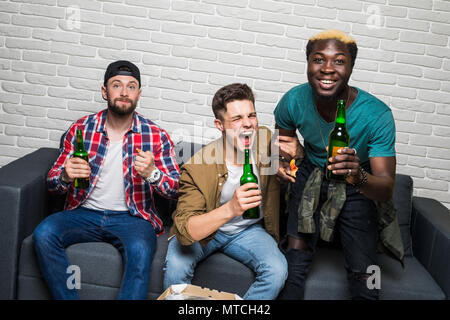 Obiettivo. Allegro giovani uomini stanno guardando la partita sul divano di casa e bere birra e mangiare la pizza. Essi sono appassionati di giochi di sport come il calcio, basket, hockey, Foto Stock