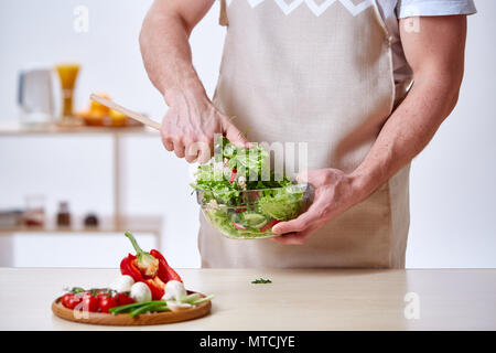 Uomo a cottura cucina facendo una sana insalata di verdure, close-up, il fuoco selettivo. Varietà di verdure sul piatto di legno sul tavolo bianco nella parte anteriore del Foto Stock