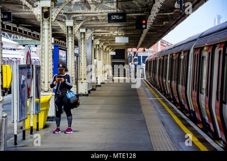 Una giovane donna sta da solo e controlla il suo telefono mobile sulla piattaforma a Hammersmith Stazione della metropolitana di Londra. Foto Stock