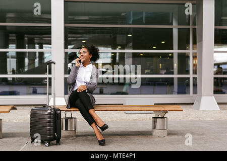 Giovane imprenditrice seduta su una panchina e parlando sullo smartphone al terminal aeroportuale Foto Stock