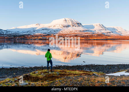 Uomo in piedi sulla riva, acqua riflessione delle montagne coperte di neve in un fiordo, Reyðarfjörður, Est Islanda Islanda Foto Stock