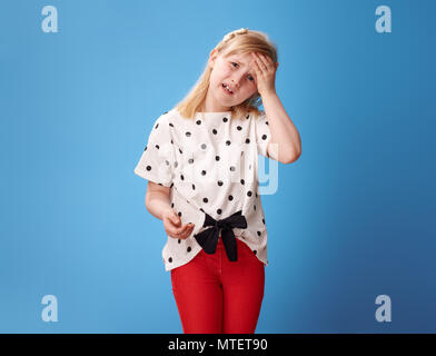 Infelice bambino moderno in pantaloni rossi aventi mal di testa isolata su sfondo blu Foto Stock