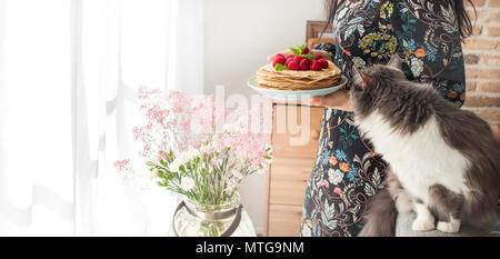 Foto di felice giovane donna in piedi presso la cucina vicino alla finestra in casa e cat. Focus sulle frittelle. Spazio di copia Foto Stock