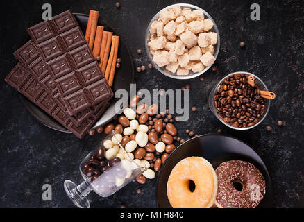 Vetro con i chicchi di caffè accanto a tavolette di cioccolato, ciambelle, lo zucchero di canna e di altro vetro con arachidi in cioccolato Foto Stock