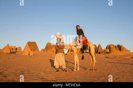 Piramidi di Meroe, Sudan- 19 dicembre 2015: una donna su un cammello con un uomo locale nella parte anteriore delle piramidi in un deserto Foto Stock