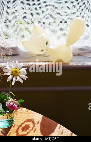 La camomilla sul vetro e il giocattolo vintage white cat sul davanzale della finestra Foto Stock