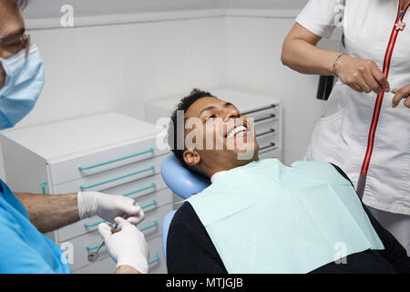 Ridendo afro-americano di uomo seduto in poltrona del dentista in clinica e preparazione per la procedura con un'infermiera. Foto Stock
