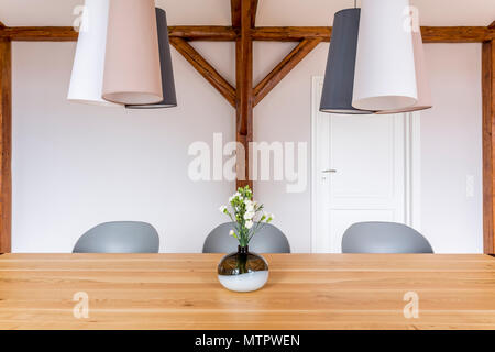 Set simmetrica di lampade e sedie grigio al tavolo da pranzo in legno con fiori di colore bianco in vaso decorativo Foto Stock
