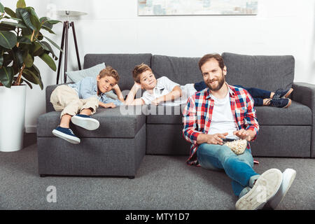Carino piccoli fratelli sdraiato sul divano mentre sorridente padre mangiare popcorn a casa Foto Stock