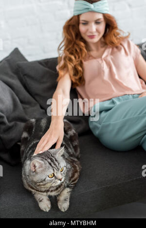 Bella giovane donna petting tabby cat comodamente seduti sul divano di casa Foto Stock