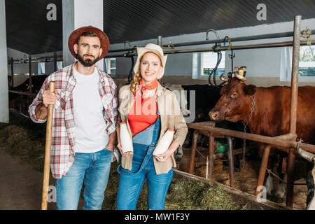 Due agricoltori con forche e bottiglie di latte fresco sorridente in telecamera in stallo Foto Stock