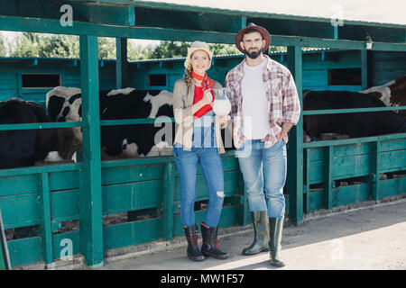 Due giovani agricoltori azienda caraffa di vetro con latte fresco e sorridente in telecamera permanente, mentre in stallo Foto Stock