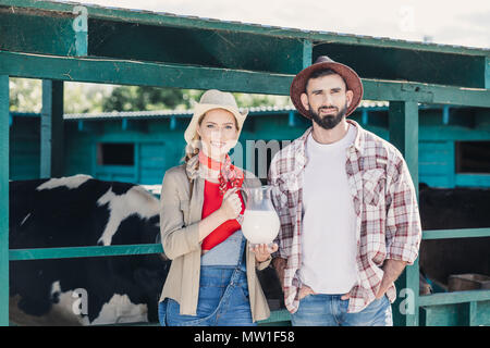 Due giovani agricoltori azienda caraffa di vetro con latte fresco e sorridente in telecamera permanente, mentre in stallo Foto Stock