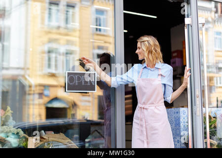 Sorridente giovane fioraio in aorn tenendo aperte segno permanente, mentre nella porta del negozio di fiori Foto Stock