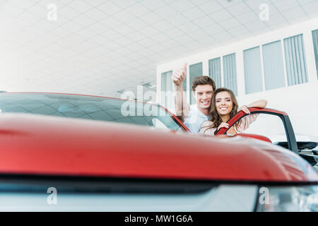 Il mio ragazzo in piedi con una ragazza in un auto showroom e mostrando il pollice fino a auto rossa Foto Stock