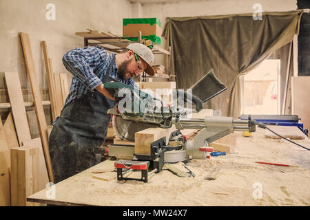 Falegname esperto in abiti da lavoro e piccole buiness proprietario lavorando nel laboratorio di falegnameria, utilizzando una sega circolare per tagliare attraverso un tavolato in legno, sul Foto Stock