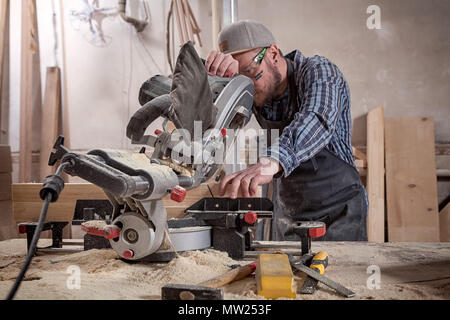 Falegname esperto in abiti da lavoro e piccole buiness proprietario lavorando nel laboratorio di falegnameria, utilizzando una sega circolare per tagliare attraverso un tavolato in legno, sul Foto Stock