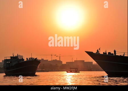 Tramonto, Dubai Creek. Grandi barche da pesca stagliano contro un bel cielo arancione con le gru e gli edifici dello skyline Foto Stock