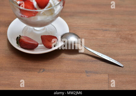 Gelato con le fragole in un recipiente di vetro su un piattino bianco con un cucchiaino e metà delle fragole. Stare al buio su un tavolo di legno Foto Stock
