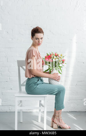 Vista laterale della bella giovane donna azienda vaso di fiori mentre è seduto su una sedia di fronte bianco muro di mattoni Foto Stock