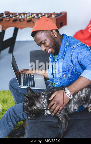 Bello African American uomo seduto sul divano con il computer portatile e accarezzare bulldog francese Foto Stock