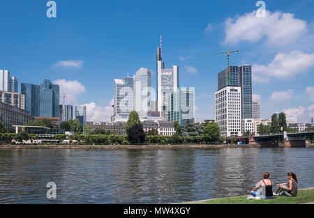 Due giovani donne seduti al sole sulla riva opposta architettura moderna dello skyline della città, del fiume Main, Frankfurt am Main, Hesse Darmstadt, Germania Foto Stock